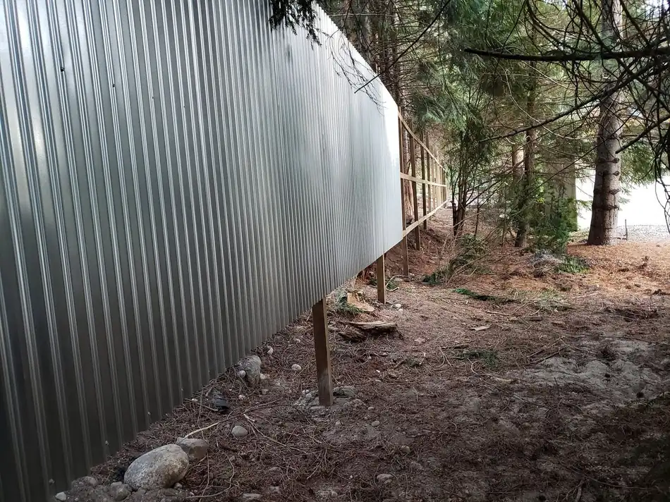 Silver corrugated metal fence line running along a wooded property boundary